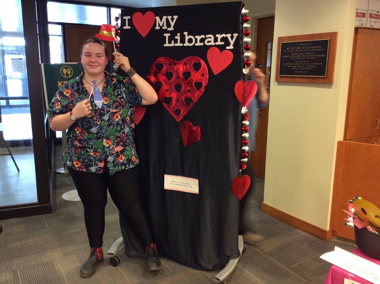 A smiling student stands in front of the I love my Library backdrop holding a prop red hat on a stick and a prop necktie on a stick up as though she is wearing them